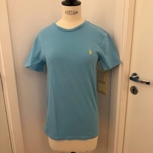 Blå / turkos t-shirt från Polo Ralph Lauren - Snygg ljusblå t-shirt från Polo Ralph Lauren med klassisk gul broderad logga på bröstet. Slim fit. Mjuk bomullskvalitet. Storlek Small. Tröjan sitter på en dam-docka men är herr stl. Fast vem som helst kan ha den såklart. Ett pytte märke i halsringningen som jag först inte såg. Loggan i nacken lite borttvättad. De två ”felen” gör att jag sätter lågt pris för en RL. Annars jättebra skick! Hämtas gärna på Hägerstensåsen eller skickas. 