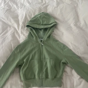 Grön croppad hoodie med dragkedja - Snygg ljusgrön hoodie med dragkedja framtill och huva. Modellen är croppad och har ribbade muddar samt en stor ficka på magen. Perfekt för en chill och avslappnad stil.