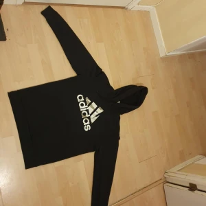 Svart Adidas hoodie med logga - Svart hoodie från Adidas med stor vit logga på bröstet. Klassisk känguruficka framtill, huva med snörning och långa ärmar. Skön passform och perfekt för chill eller träning. Materialet är mjuk bomullsmix som känns nice mot huden.