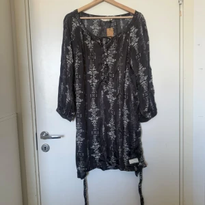 Mörkgrå mönstrad kortklänning från Hunkydory - Mörkgrå kortklänning från Odd Molly med vitt blommigt mönster. Klänningen har trekvartsärmar, snörning framtill och är tillverkad i ett tunt, luftigt material. Perfekt för dig som gillar boho-vibe och avslappnad stil. Storlek 2 som enligt deras storleksguide motsvarar en storlek M.  Mycket fint skick, jag hittar inga synliga anmärkningar eller defekter.