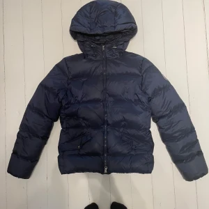 Mörkblå dunjacka från Gant med huva - Snygg mörkblå dunjacka från Gant med huva och dragkedja framtill. Jackan har två fickor med lock och en klassisk pufferstil. Perfekt för kalla dagar och riktigt skön att bära. Insidan har Gant-text och röda detaljer längs fodret. OBS!!! Det kan finnas pyttesmå vita fläckar INUTI jackan som säkert går att få bort. Det är skrivet på i lappen på sidan innanpå jackan. Originalpris ca 3000