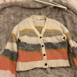 Randig kofta från Name It i stickad bomull - Supermysig stickad kofta från Name It med breda ränder i beige, grått, rosa, gult och vitt. Koftan har v-ringning, långa ärmar och knappar framtill. 