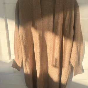 Beige kofta från H&M i storlek M - Mysig beige kofta från H&M i storlek M. Koftan är lång, har långa ärmar och är stickad i ett mjukt material. Perfekt att slänga på sig när det är kyligt ute. Enkel och stilren design utan knappar eller dragkedja.
