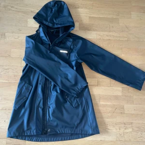 Mörkblå regnjacka True North S - Snygg mörkblå regnjacka från True North i storlek S. Jackan har huva, långa ärmar, två fickor med lock och både dragkedja och knappar framtill. Perfekt för blöta dagar och har en clean, stilren look som funkar till många outfits. Pris kan diskuteras. 👍 