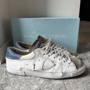 Philippe Model sneakers - Snygga vita sneakers från Philippe Model med grå mockadetaljer på tån och blå häl. Skorna har klassisk snörning och en rund tå. Sulan är svart med stora bokstäver i beige. Tillverkade i skinn och mocka för en cool och urban vibe.