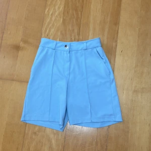 Ljusblåa högmidjade shorts - Snygga ljusblåa shorts med hög midja och klassisk knappstängning. De har raka ben, pressveck framtill och sidofickor för en clean look. Perfekta för varma dagar när du vill ha en stilren vibe.