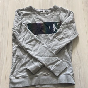 Grå Calvin Klein Jeans sweatshirt - Snygg grå sweatshirt från Calvin Klein Jeans med stort CK-logga tryck på bröstet i mörka nyanser. Tröjan har rund hals, långa ärmar och ribbade muddar. Perfekt för en chill och stilren look. Priset är ej hugget i sten. Tveka inte att ställa frågor vid minsta fundering.