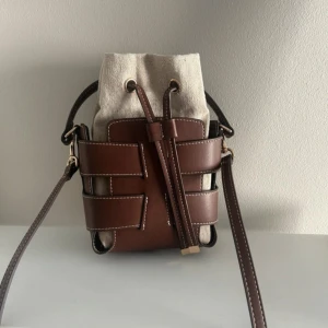 Brun och beige bucket bag med snörning - Säljer en snygg bucket bag i brun skinnimitation och beige tyg. Väskan har coolt flätat mönster i skinn och stängs med snörning upptill. Justerbar axelrem och handtag gör den enkel att bära. Perfekt storlek för det viktigaste och ger en trendig touch till din outfit.