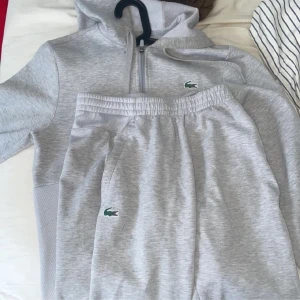 Grå mjukisbyxor från Lacoste sport - Snygga ljusgrå mjukisbyxor från Lacoste med klassisk krokodillogga på benet. Byxorna har resår i midjan och är tillverkade i mjukt bomullsmaterial för maximal komfort. Perfekta för chill dagar eller när du vill ha en sportig look. Koftan är XS byxor S