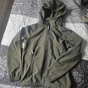 Grön vindjacka från Kayoba Outdoor - Snygg grön vindjacka från Kayoba Outdoor med huva och hel dragkedja framtill. Jackan har två fickor med dragkedja, elastiska muddar och vita detaljer längs sömmarna. Perfekt för uteliv och äventyr, stilren och funktionell design.