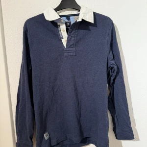 Randig pikétröja från Hansen & Jacob - Snygg långärmad pikétröja från Hansen & Jacob i blått och beige med breda ränder på ryggen. Vit krage, knappar framtill och quiltade armbågslappar. Broderad logga på ärmen och detaljerad insida på kragen. 