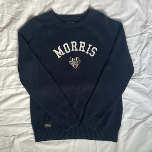 Mörkblå stickad tröja Morris - Snygg mörkblå stickad tröja från Morris med vit broderad logga och emblem på bröstet. Rund hals och ribbade muddar vid ärmslut och nederkant. Perfekt för dig som gillar klassisk college-stil och vill ha något som funkar till jeans eller chinos.