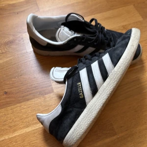Adidas Gazelle svarta sneakers mocka -  Storlkek 41 ganska dåligt skick därför endast 399kr. Skorna har platt sula, rund tå och snörning. Guldtexten 'GAZELLE' på sidan och vit hälkappa med Adidas-logga. Perfekta för en clean och sportig stil. Skriv vid minsta fundering.