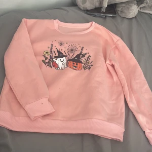 Rosa sweatshirt med halloweenmotiv - Supermysig rosa sweatshirt med tryck av ett gulligt spöke och en pumpa med häxhattar bland blommor. Tröjan har långa ärmar, ribbade muddar och insida i mjuk fleece. Perfekt för dig som älskar höst och halloween!