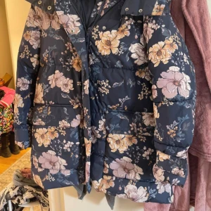 Blommig pufferjacka från House Brand XS - Supersnygg pufferjacka från House Brand i storlek XS. Jackan är mörkblå med stora blommiga mönster i beige, gult och ljusrosa. Den har huva, stora fickor framtill och stängs med dragkedja och knappar. Perfekt för att sticka ut under kalla dagar! Rymlig i storlek. Sparsamt använd. 
