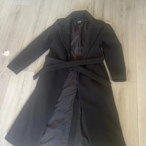 Svart kappa med bälte från ONLY - Säljer en svart lång kappa från ONLY i storlek M. Klassisk modell med bred krage, knytbälte i midjan och helfodrad insida. Perfekt för dig som vill ha en stilren och tidlös look under höst och vinter. kostade 1200kr