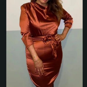 Kopparfärgad satin klänning med knyt - Snygg kopparfärgad kort klänning i satin med 3/4-ärmar och ett matchande knytband i midjan. Klänningen har en rund halsringning och figurnära passform som framhäver siluetten. Perfekt för dig som gillar glansiga material och vill sticka ut.