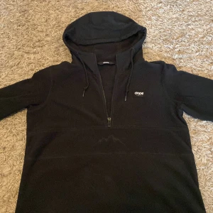 Svart fleece hoodie från Dope - Svart hoodie från Dope med halv dragkedja och broderat bergsmotiv på framsidan. är gjord i mjuk fleece som håller dig varm. Perfekt för kalla dagar och chill stunder. Snygg logga på bröstet och stilren design.