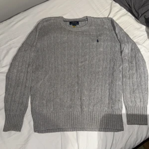 Grå kabelstickad tröja Polo Ralph Lauren - Snygg grå kabelstickad tröja från Polo Ralph Lauren med klassisk rund hals och mörkblå broderad logga på bröstet. Tröjan har långa ärmar och ribbade muddar vid ärmslut och nederkant. Perfekt för dig som gillar stilren och tidlös design.