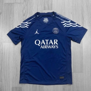 PSG blå matchtröja D. Doué #14 - Snygg PSG fotbollströja i mörkblått med vita detaljer och tryck. Jordan-logga på bröstet, Qatar Airways sponsor och PSG-emblem. Unikt mönster på axlarna, Champions League-märke på ärmen och D. Doué #14 på ryggen. Tillverkad i lätt och ventilerande material.