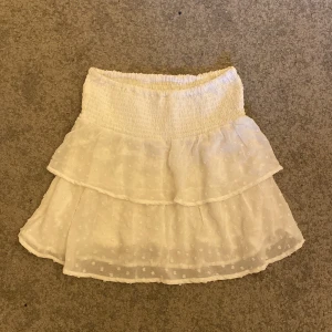 Vit volangkjol med prickar - Supersöt vit kortkjol med volanger och diskret prickigt mönster. Kjolen har smock i midjan för en bekväm passform och är gjord i ett lätt, luftigt tyg som passar perfekt till varma dagar.