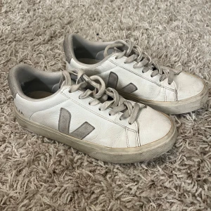 Vita Veja sneakers  - Snygga vita Veja sneakers med grå detaljer och logga på sidan. Skorna har snörning, rund tå och platt sula. Ovandelen är i skinn och hälen samt V-loggan är i mocka. Perfekta för en clean och stilren look.