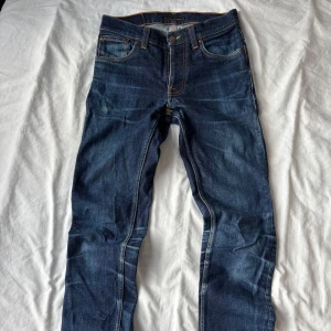 Blå jeans från Nudie Jeans Co - Snygga blå jeans från Nudie Jeans Co med klassisk femficksdesign och orangea kontrastsömmar. Jeansen är tillverkade i ekologisk bomull och har en mörk tvätt med lätt slitning. Bakfickan har Nudie Jeans signaturbrodyr. Perfekta för en avslappnad stil. Modellen är slim.