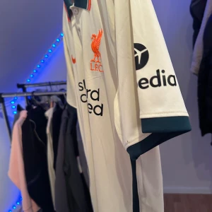 Liverpool FC Nike vit bortatröja - Liverpool FC bortatröja från Nike i vit polyester med broderat rött klubbmärke och Nike-logga. Tröjan har korta ärmar med mörkgröna muddar och klassisk krage med röda detaljer. Snygg och sportig design med tryckta sponsorer på bröst och ärm.