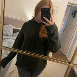 Mörkgrå stickad hoodie med huva - Mysig mörkgrå stickad hoodie med huva och snörning. Tröjan har en loose passform och långa ärmar med ribbade muddar. Perfekt för chill dagar och när du vill ha det extra bekvämt. Snygg att matcha med jeans eller leggings.