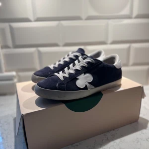 The gallerist skor - Snygga mörkblå sneakers från The Gallerist med vit snörning och vit detalj i form av en klöver på sidan. Skorna har vit häl med logotyp och mönstrad remsa bak. Ovandelen är i mocka och insidan är fodrad med vitt material. Perfekt för dig som gillar unik design.Riktigt fint skick!