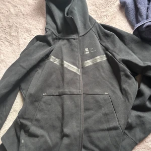 Svart Nike tech hoodie med dragkedja - Svart hoodie från Nike med hel dragkedja och huva. Tröjan har två stora fickor framtill och diskreta gråa ränder över bröstet. Perfekt för chill dagar eller när du vill ha en sportig look. Klassisk Nike-logga på bröstet och mjukt material.