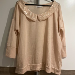 Beige stickad tröja med paljettkrage - Supersöt beige stickad tröja från ONLY med bred ribbad kant och trekvartsärmar. Kragen är dekorerad med paljetter och spetsdetaljer som ger en extra touch. 