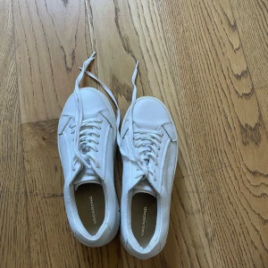 Vita sneakers från Vagabond - Säljer ett par fräscha vita sneakers från Vagabond i storlek 37. Skorna har klassisk rund tå, platt sula och snygga detaljer i vitt skinn. De har snörning och en stilren design som passar till många olika outfits. Perfekta för dig som gillar minimalistisk stil.