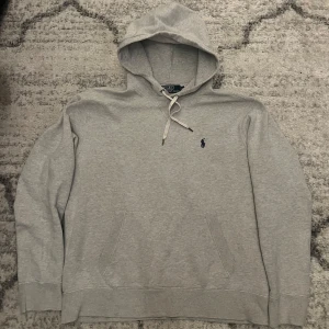 Grå hoodie från Polo Ralph Lauren - Snygg grå hoodie från Polo Ralph Lauren med klassisk logga broderad i mörkblått på bröstet. Tröjan har huva med snörning, känguruficka och långa ärmar. Perfekt för chill dagar och enkel att matcha med jeans eller joggers.