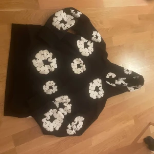 Svart hoodie Denim Tears blommig  - Svart hoodie från Denim Tears med stora vita blommönster över hela tröjan och huva. Snygg och unik streetwear vibe, perfekt för dig som vill sticka ut. Tillverkad i USA och har en avslappnad passform med långa ärmar.