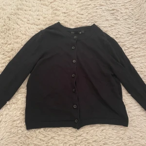 Svart kofta från lager 157  - En klassisk svart kofta från 15% med rund halsringning och knäppning framtill. Långärmad modell i mjukt material, perfekt att slänga över en t-shirt eller linne. Ribbstickade muddar vid ärmslut och nederkant ger en clean look.