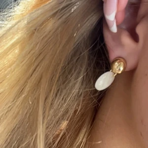 Guldiga örhängen med vit marmordetalj - Snygga örhängen med rund, blank guldplatta upptill och hängande vit marmorerad cirkel i plast. De har push-back stängning och en modern, stilren look som passar till många outfits. Perfekt för dig som gillar trendiga accessoarer med lite extra edge.