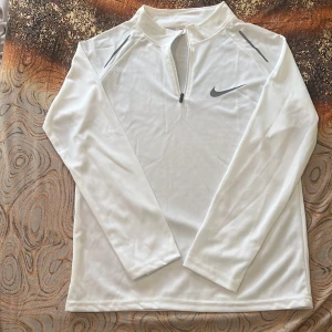 Vit långärmad Nike half zip tröja - Snygg vit långärmad tröja från Nike med halv dragkedja framtill och gråa detaljer på axlarna. Perfekt för träning eller chill dagar. Tillverkad i mjukt och lätt polyestermaterial som andas. Klassisk Nike Swoosh på bröstet.