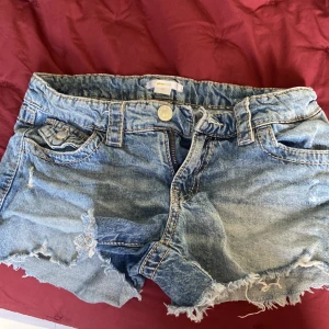Blå jeansshorts från Gina Tricot - Snygga blå jeansshorts från Gina Tricot med slitna detaljer och fransig kant. Klassisk femficksmodell med knappar och synliga sömmar i kontrastfärg. Perfekta för varma dagar och en avslappnad look.