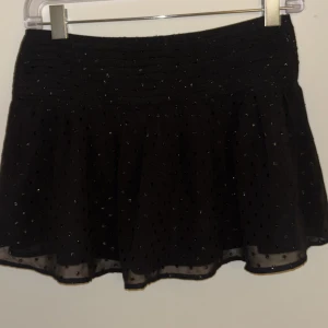 Svart glittrig kortkjol Gina Tricot - Supersöt svart kortkjol från Gina Tricot med glittriga detaljer och lätt volang. Kjolen har ett smockat midjeparti och små glittrande prickar över hela tyget. Perfekt för dig som vill sticka ut lite extra!