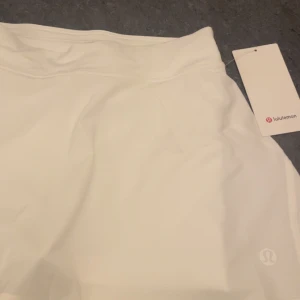 Vit tennis kjol från Lululemon - Oanvänd, Superfräsch vit kortkjol från Lululemon med hög midja och inbyggda shorts. Kjolen är tillverkad i lätt och snabbtorkande polyesterblandning med fyrvägsstretch för maximal rörelsefrihet. Perfekt för tennis eller träning, stilren och cute look.