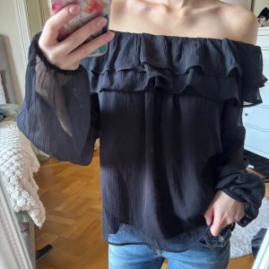 Svart offshoulder blus med volang - Säljer en svart offshoulder blus i luftig polyester med volangdetaljer upptill och långa, transparenta ärmar. Blusen är från Gina Young i st 158/164 men passar xs och nedåt. 