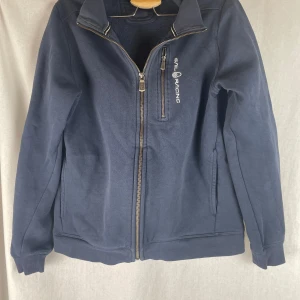 Marinblå jacka från Sail Racing - Snygg marinblå jacka från Sail Racing med dragkedja framtill och hög krage. Jackan har en bröstficka med dragkedja och logga på bröstet. Tillverkad i mjuk bomullsmix, perfekt för vår och höst. Långärmad och stilren design som funkar till det mesta.