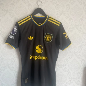Manchester United 25/26 matchtröja Amad 16 - Snygg svart Manchester United fotbollströja från Adidas med gula detaljer, klubbmärke och sponsortryck. Tröjan har gula ränder på axlarna, blå och gula detaljer vid krage och ärmslut samt namn och nummer Amad 16 på ryggen.