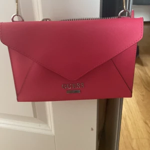 Rosa kuvertväska från Guess - Snygg rosa kuvertväska från Guess med silverfärgade detaljer och dragkedja upptill. Väskan har ett stilrent kuvertformat lock och flera fack inuti för kort och småsaker. Tillverkad i syntetmaterial och perfekt att bära crossbody med kedjerem. Helt ny. Helt nytt pris är 1200kr