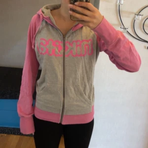 Grå och rosa zip-up från Nikita - Grå zip-up hoodie från Nikita med rosa ärmar, huva och muddar. Dragkedja framtill och stor rosa logga på bröstet. Skön och avslappnad passform. Tillverkad i mjuk bomullsmix och har praktiska fickor framtill.