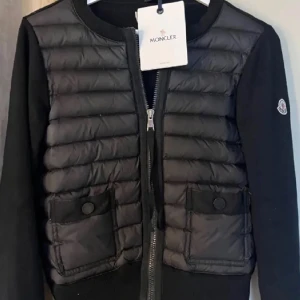 Moncler cardigan - Snygg svart dunväst från Moncler med quiltad framsida, två fickor med knapp och dragkedja framtill. Västen har Moncler-logga på ärmen och är i en croppad modell som ger en trendig look. Perfekt att styla över en hoodie eller långärmad tröja.