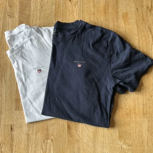 2-pack GANT t-shirts vit & marinblå - Två klassiska t-shirts från GANT i storlek 146/152. En är vit och en är marinblå, båda med GANT-logga och sköld på bröstet. T-shirtsen är i mjuk bomull och har rund hals och korta ärmar. Perfekta för en clean och stilren look.