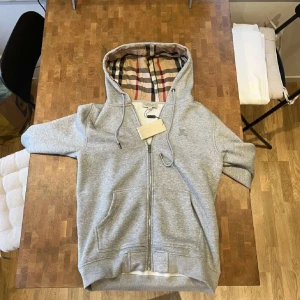 Grå hoodie med dragkedja från Burberry - Säljer en grå hoodie med dragkedja från Burberry. Insidan av huvan har det klassiska rutiga Burberry-mönstret i beige, svart och rött. Tröjan har snörning i huvan och två fickor framtill. Perfekt för en stilren och avslappnad look.