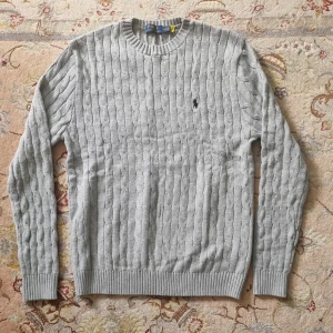 Grå kabelstickad tröja Polo Ralph Lauren - Snygg grå kabelstickad tröja från Polo Ralph Lauren med klassisk rund halsringning och broderad logga på bröstet. Tröjan har ribbade muddar och är perfekt för lager-på-lager-stil under kyliga dagar. Självklart äkta. Priset kan diskuteras vid snabb och smidig affär 😊